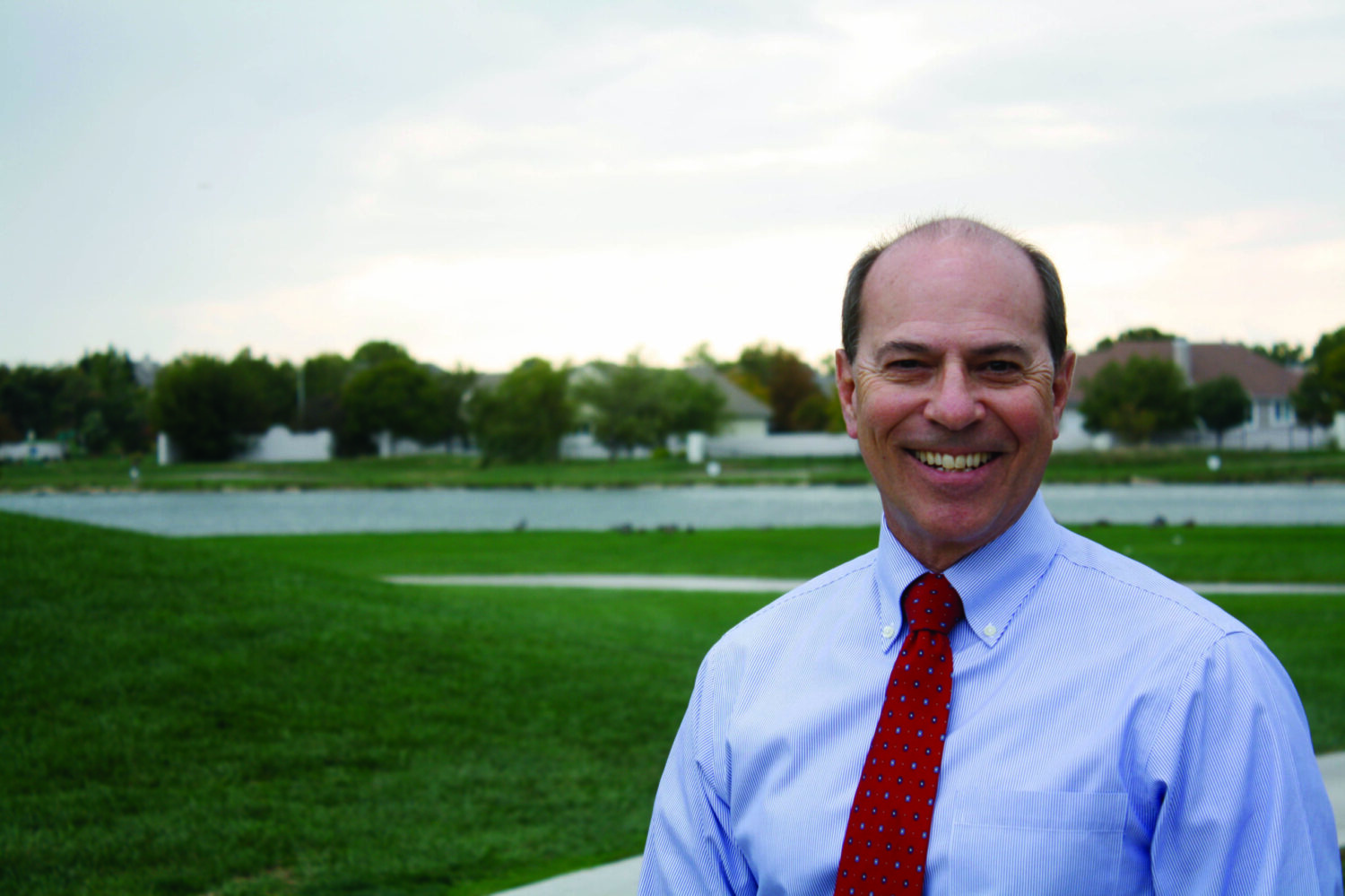 White man in blue shirt and red tie standing in front of a golf course backdrop. He is smiling.