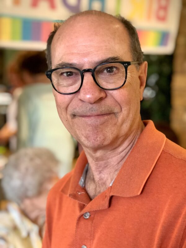 White man with black glasses and balding hair smiling at the camera. He is wearing an orange polo shirt.